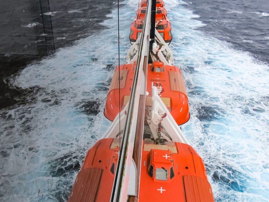Orange lifeboats lined along the side of a ship moving through blue water
